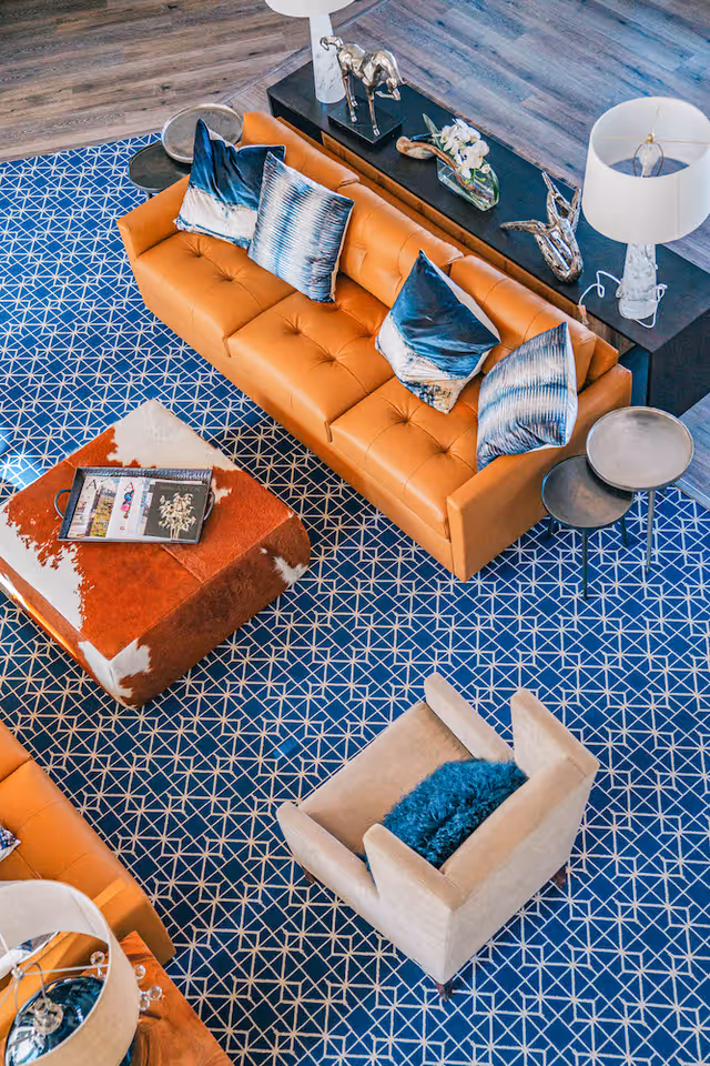 Aerial view of a stylish seating area with a tan leather sofa, patterned blue rug, cowhide ottoman, and beige armchair.