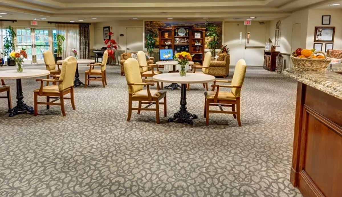 A spacious and well-lit common area with round tables and cushioned chairs arranged on a patterned carpet. There are flower vases on the tables, a wooden bookshelf with decorative items and plants in the background, and a granite countertop with a basket of fruit on the right side. Large windows allow natural light to enter the room.