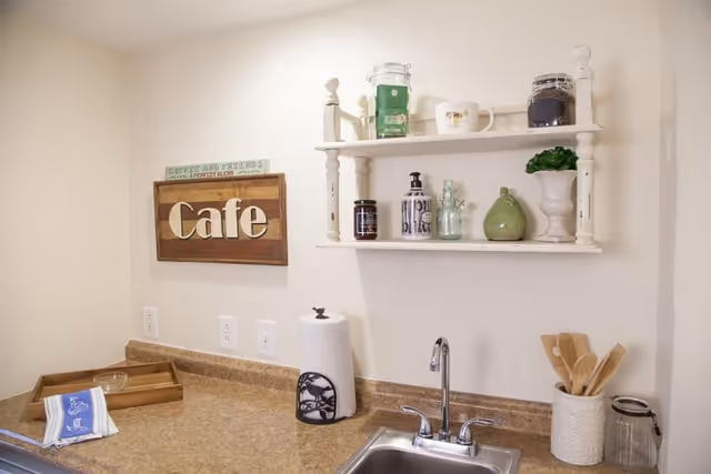 A small kitchen area with a countertop, a sink with a faucet, a paper towel holder, and a container holding wooden utensils. Above the countertop is a white wall shelf displaying jars, a cup, and a small plant. A wooden sign on the wall reads 'Cafe'.