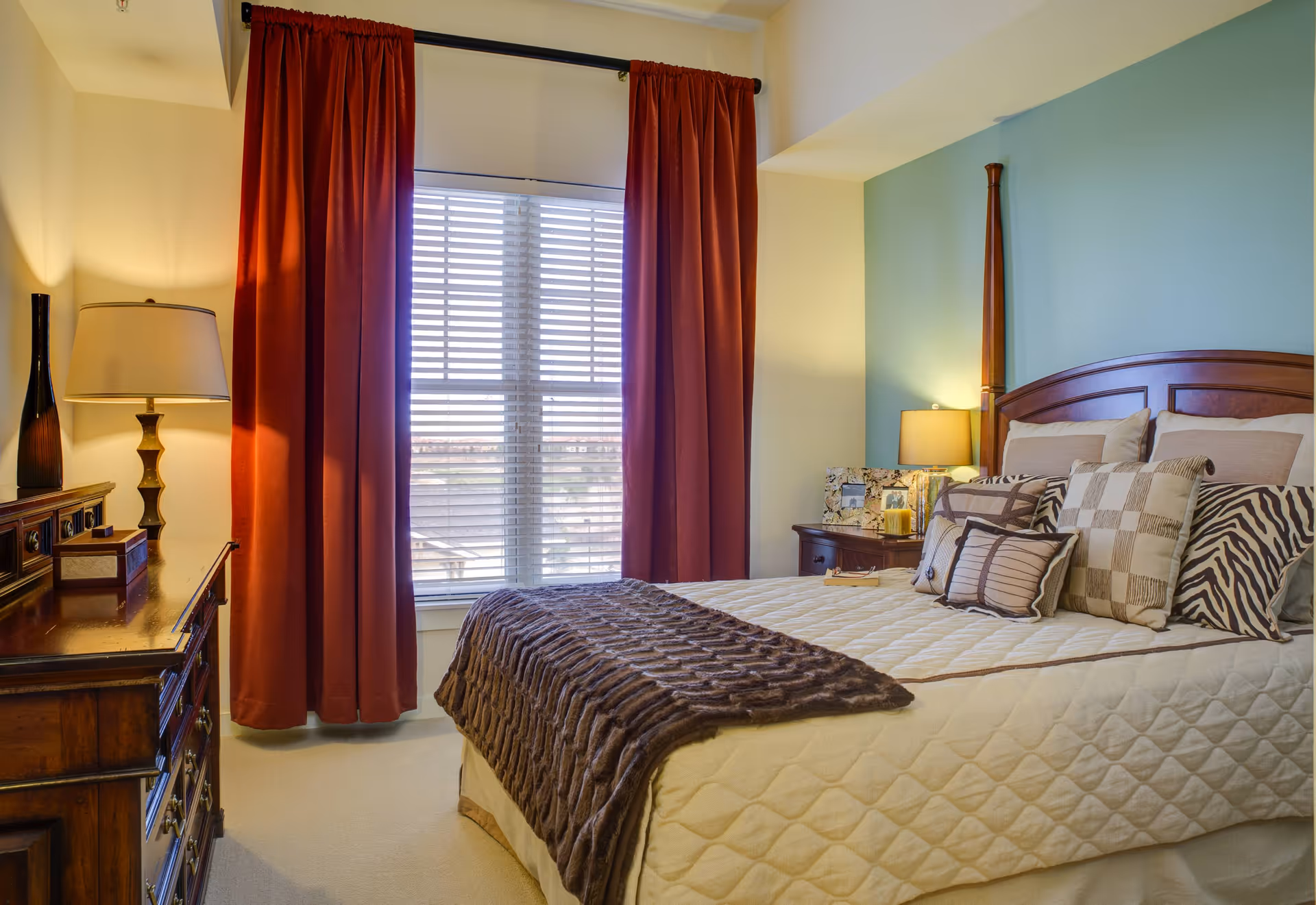 Bright bedroom with a neatly made bed, patterned pillows, nightstands with lamps, a dresser, and a window with red curtains.