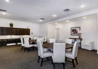 Bright communal dining room with round tables and upholstered chairs and a kitchenette along the back wall.