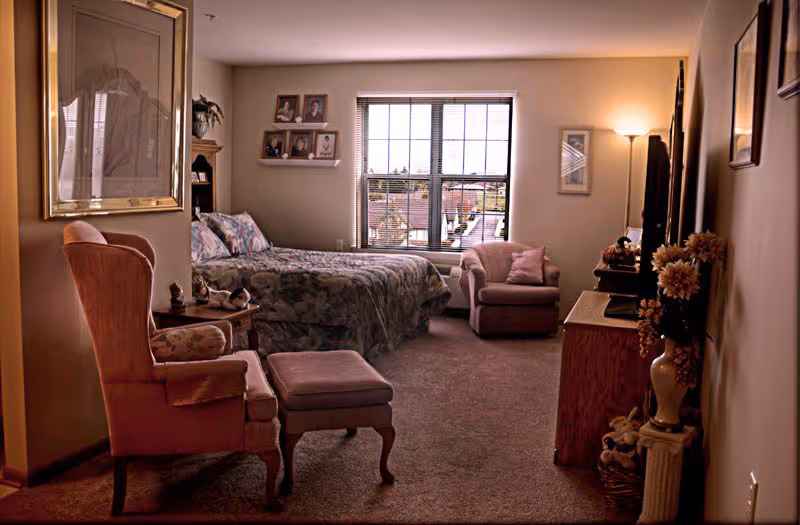 Cozy senior living bedroom with a bed, armchairs, a TV dresser, and a window overlooking the outdoors.
