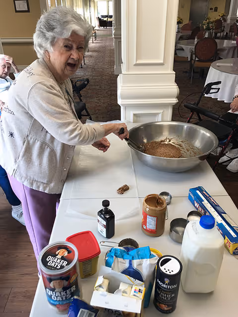 An elderly woman is standing at a table in a communal dining or activity area, stirring a large metal bowl containing a mixture. Various baking ingredients including Quaker Oats, peanut butter, vanilla extract, milk, salt, and wax paper are spread out on the table. Other elderly individuals and walkers are visible in the background.