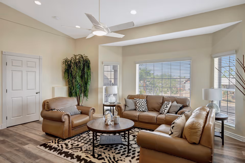 A bright living room with beige walls and large windows with blinds. The room features a brown leather sofa, two matching armchairs, a round wooden coffee table with decorative items, a patterned area rug, two side tables with lamps, a tall green plant in the corner, and a white ceiling fan with lights.