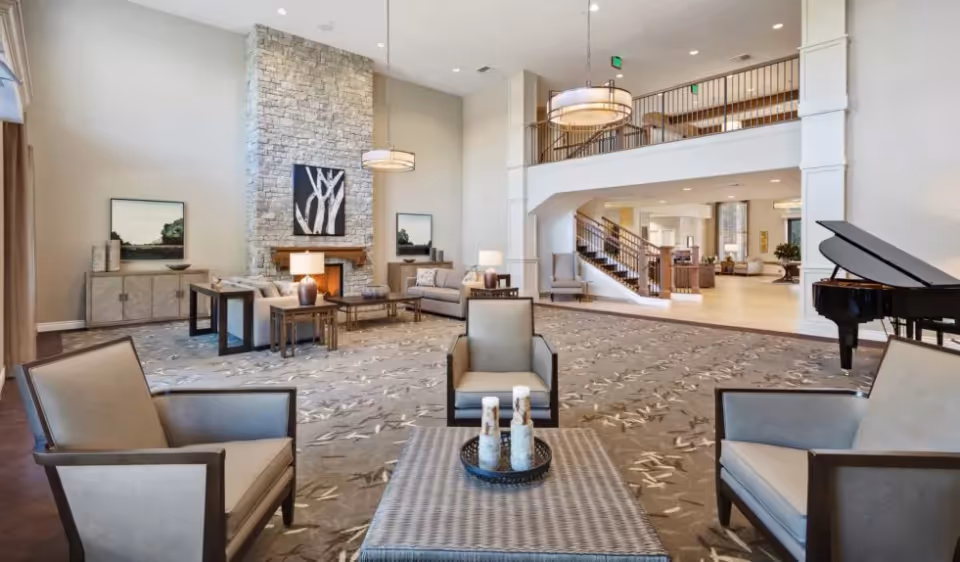 Spacious senior living community lounge with seating around a coffee table, a stone fireplace, grand piano, and a mezzanine balcony.