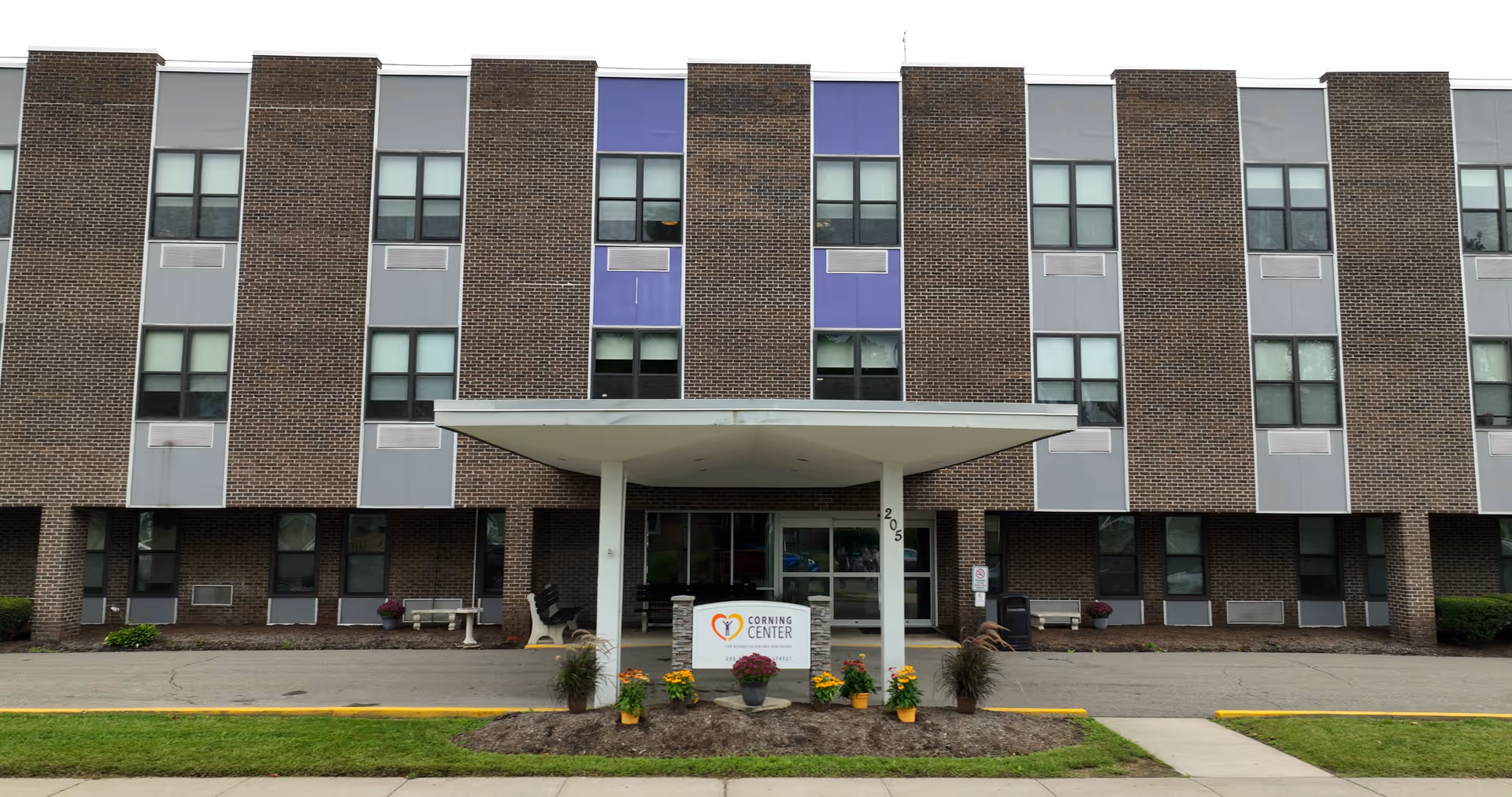 Front exterior view of the Corning Center for Rehabilitation and Nursing, a three-story brick building with multiple windows and a covered entrance. There are potted plants and flowers near the entrance sign displaying the facility's name and logo.