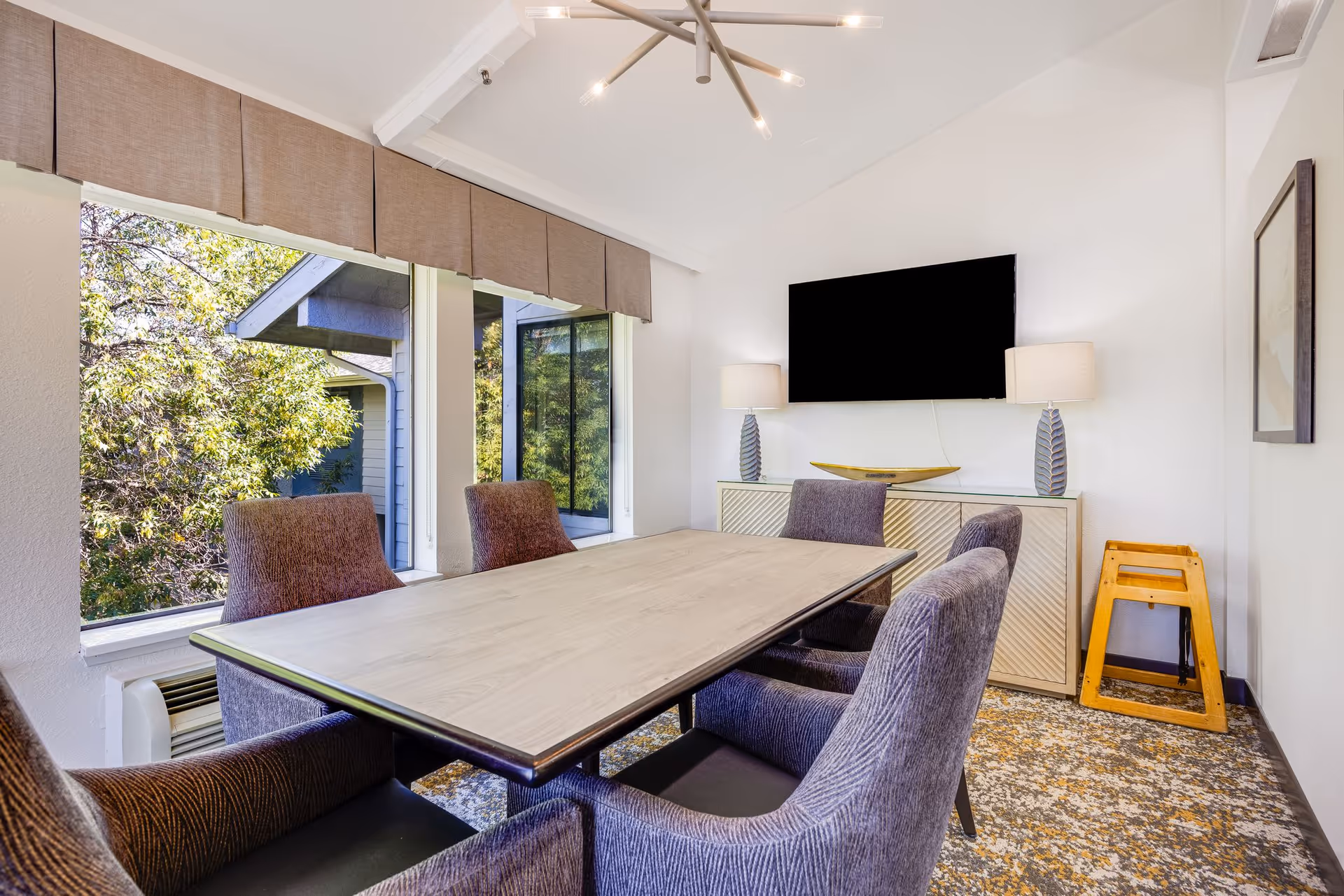 A bright meeting or dining room with a rectangular wooden table surrounded by six upholstered chairs. The room has large windows with beige valances, allowing natural light and a view of trees outside. On the far wall, there is a flat-screen TV mounted above a sideboard with two table lamps and a decorative bowl. A wooden step stool is placed in the corner.