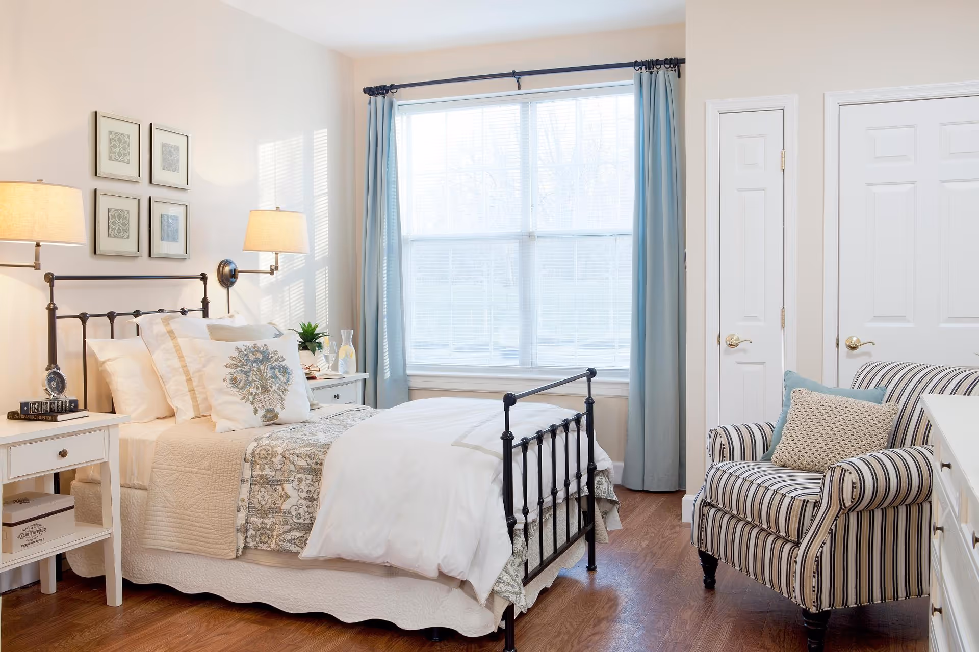 Bright furnished bedroom with an iron bed, bedside tables and lamps, a striped armchair, and a large window with light blue curtains.