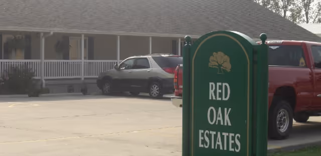 Parking lot in front of a single-story building with a porch and white railing. Several vehicles are parked, including a gray SUV and a red pickup truck. A green sign in the foreground reads 'RED OAK ESTATES' with a tree logo above the text.