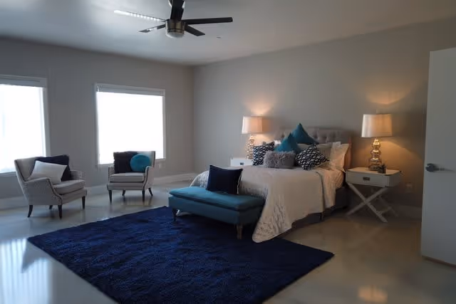 A spacious bedroom with a large bed adorned with multiple pillows, two bedside tables each with a lamp, a blue bench at the foot of the bed, two armchairs near two windows, and a dark blue rug on the floor. The room has a ceiling fan and neutral-colored walls.