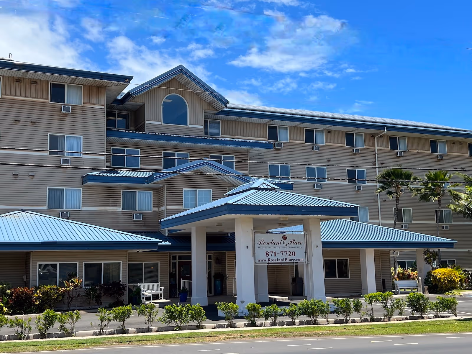 Front exterior of a four-story senior living building with blue metal roofs and a covered entrance displaying a Roselani Place sign.