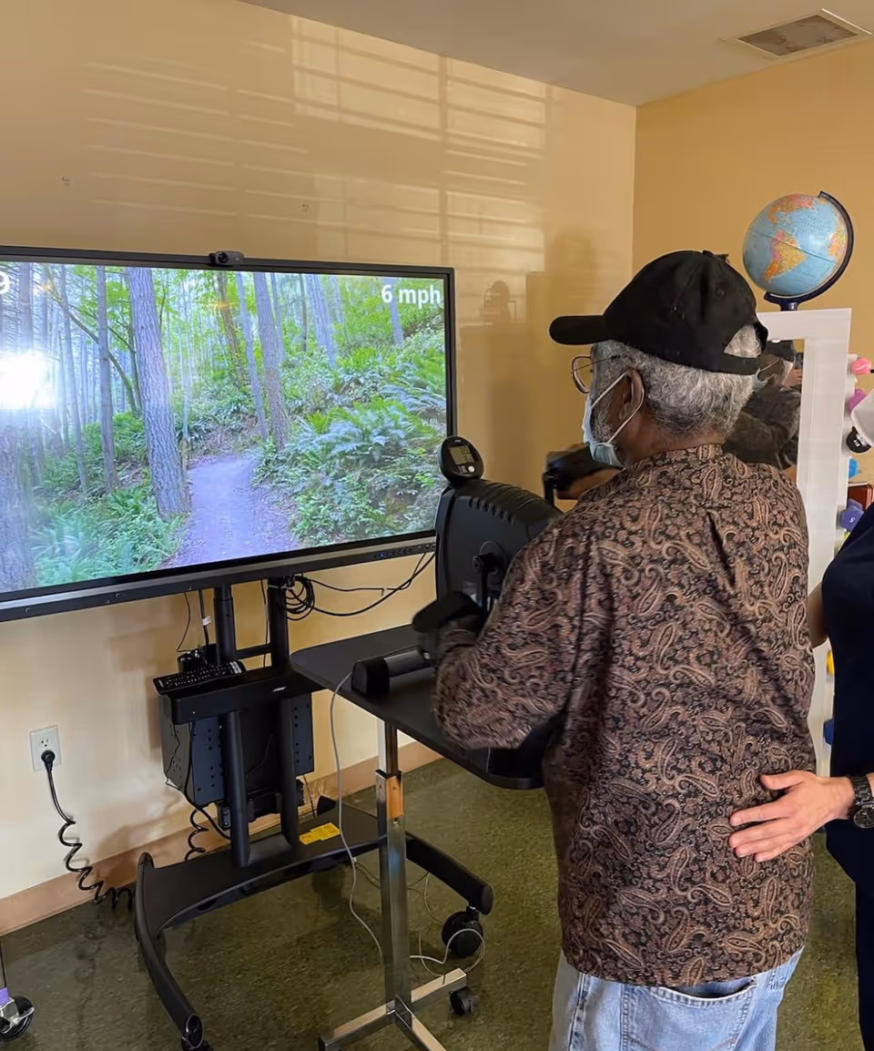 An elderly man wearing a patterned shirt, black cap, glasses, and a face mask is using a stationary exercise bike in front of a large screen displaying a forest trail with trees and greenery. A person standing beside him is gently supporting his back. The room has beige walls and a globe on a white shelf in the background.