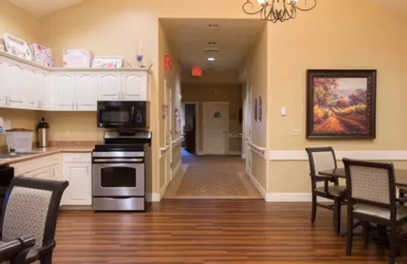 Interior view of a memory care community showing a kitchen area with white cabinets, a stainless steel stove and microwave, and a countertop with a coffee pot. To the right, there is a dining area with a table and chairs, and a framed painting on the wall. The floor is wooden, and a hallway extends in the background with beige walls and carpet.