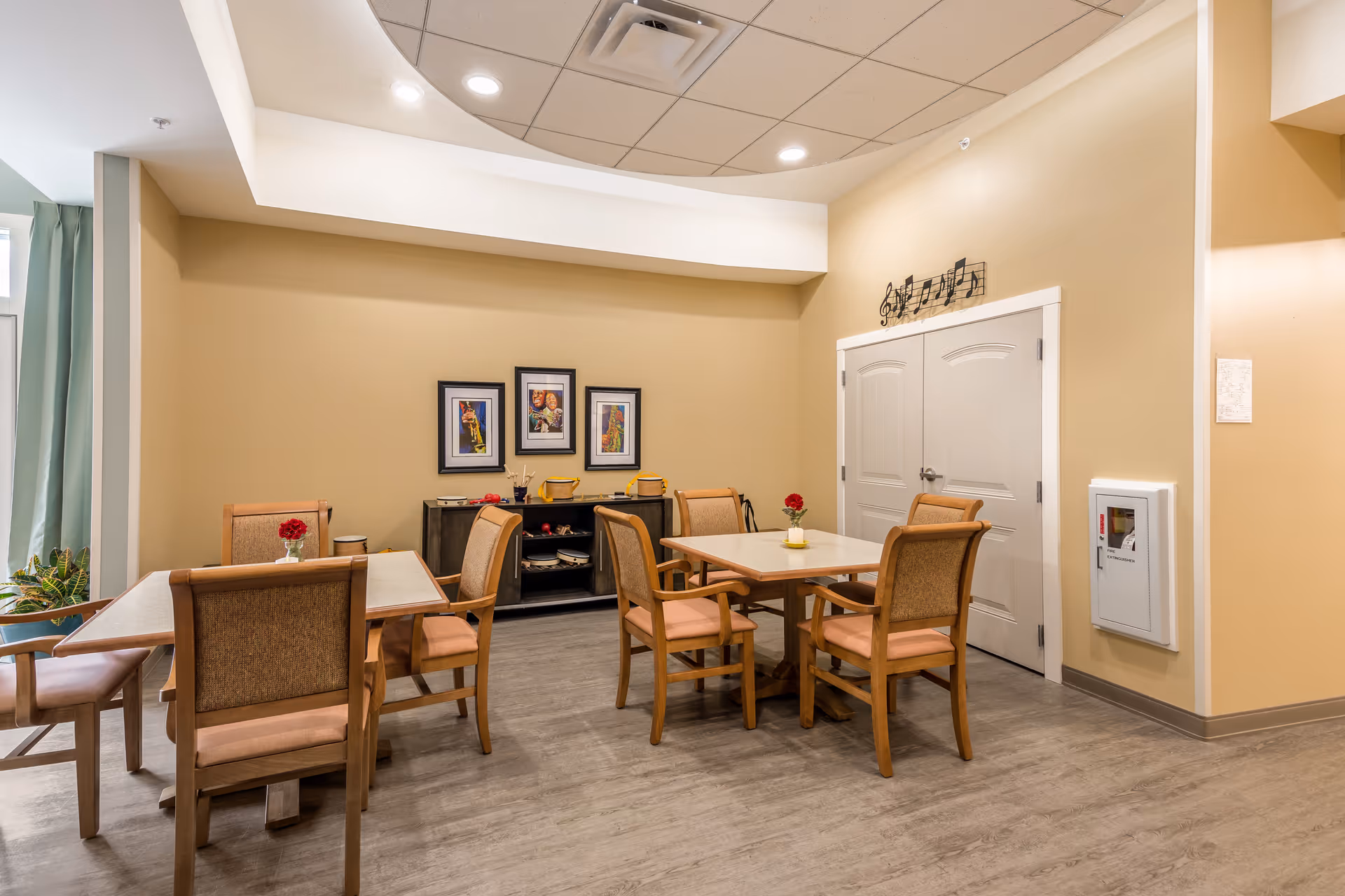A cozy dining area in a senior living facility with two square tables, each surrounded by four wooden chairs with cushioned seats. The walls are painted beige and decorated with three framed pictures and a musical note wall art above double doors. The floor is a light wood laminate, and there are small vases with red flowers on the tables.