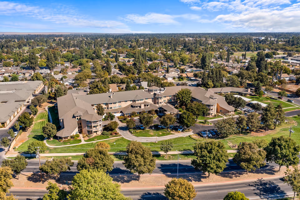 Aerial view of The Hampshire - A Provincial Senior Living Community, showing a large multi-wing building surrounded by green lawns, trees, and parking areas, with a residential neighborhood in the background under a clear sky.