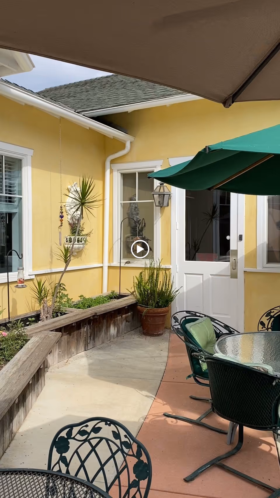 Outdoor patio area with a yellow building wall, white door, and windows. There are green metal chairs with cushions around a glass-top table, a large green umbrella providing shade, and various potted plants and garden beds along the wall.