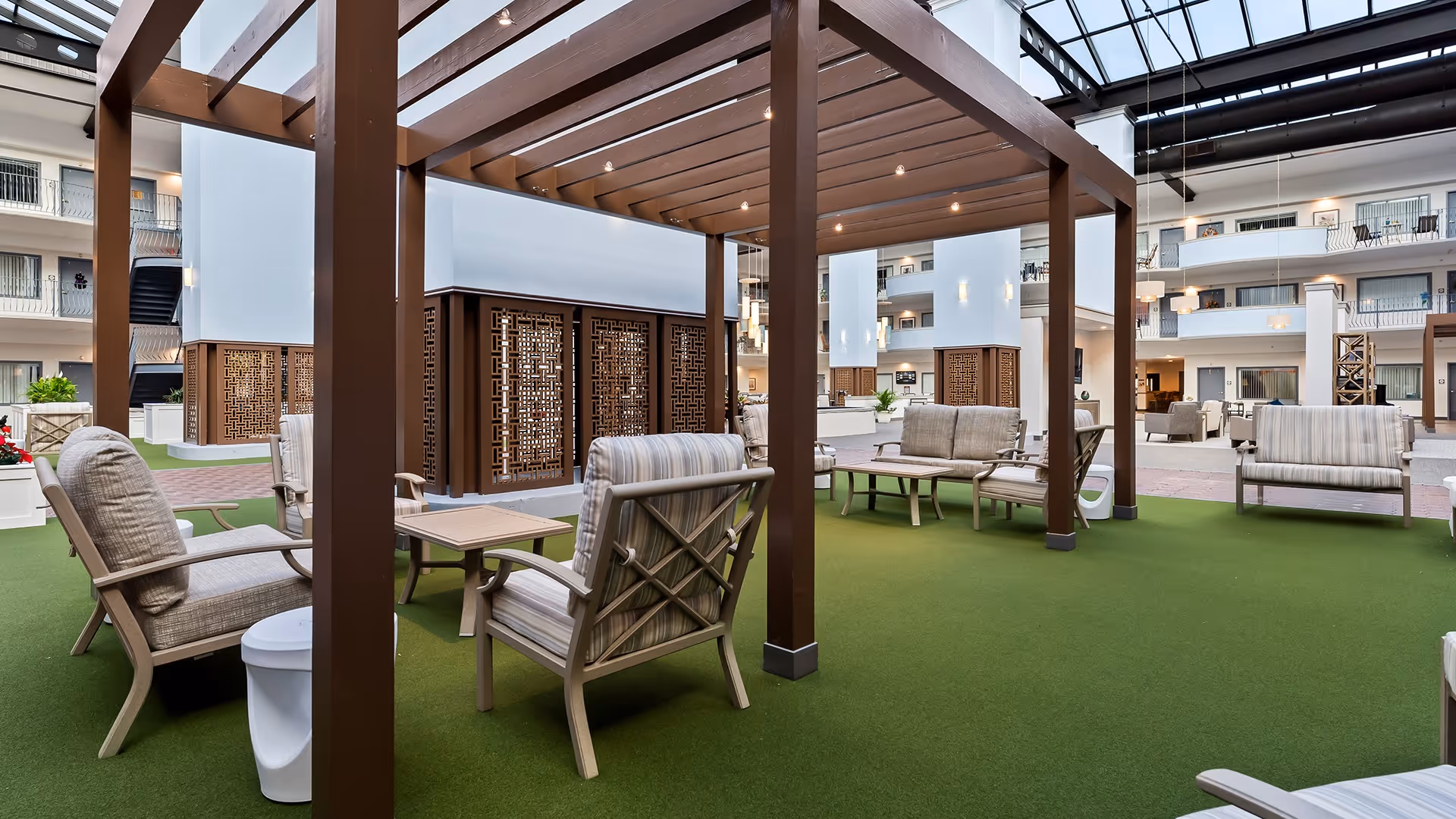 Indoor seating area at Provincial Glenville with cushioned chairs and small tables arranged under a wooden pergola structure on green carpet flooring, surrounded by a multi-story building with balconies and large windows.