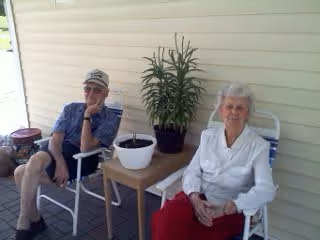 An elderly man and woman sitting on white chairs on a porch or patio area. The man is wearing a blue shirt, shorts, and a cap, while the woman is dressed in a white blouse and red pants. Between them is a small table with two potted plants. The background shows light-colored siding of the building.