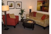 Cozy living room with a beige sofa, red armchair, oval coffee table, lamps, potted plant, and framed artwork on the walls.