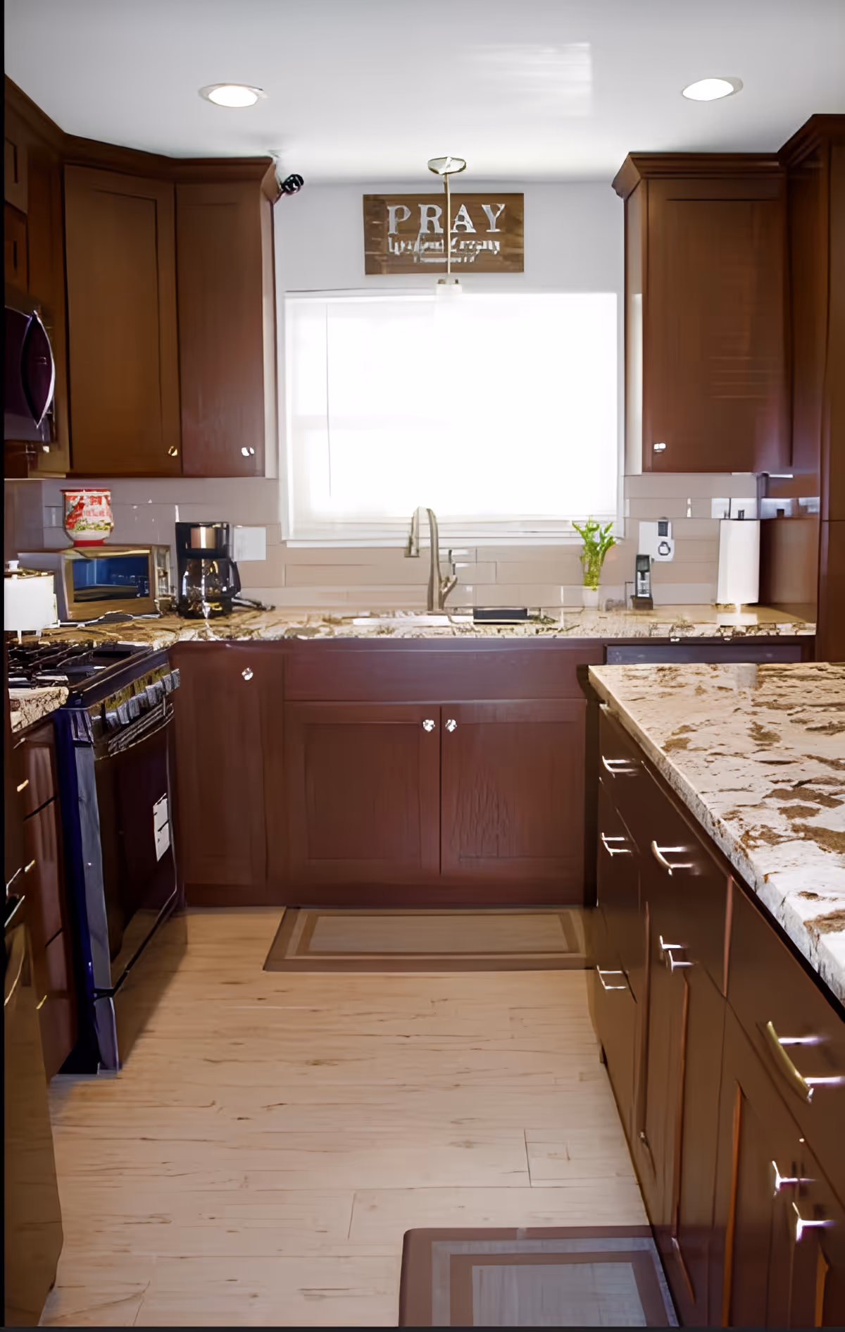 A modern kitchen with dark wooden cabinets, granite countertops, a stainless steel sink under a window with a white blind, a coffee maker, microwave, stove, and a small plant on the counter. There is a decorative sign above the window that reads 'PRAY'.