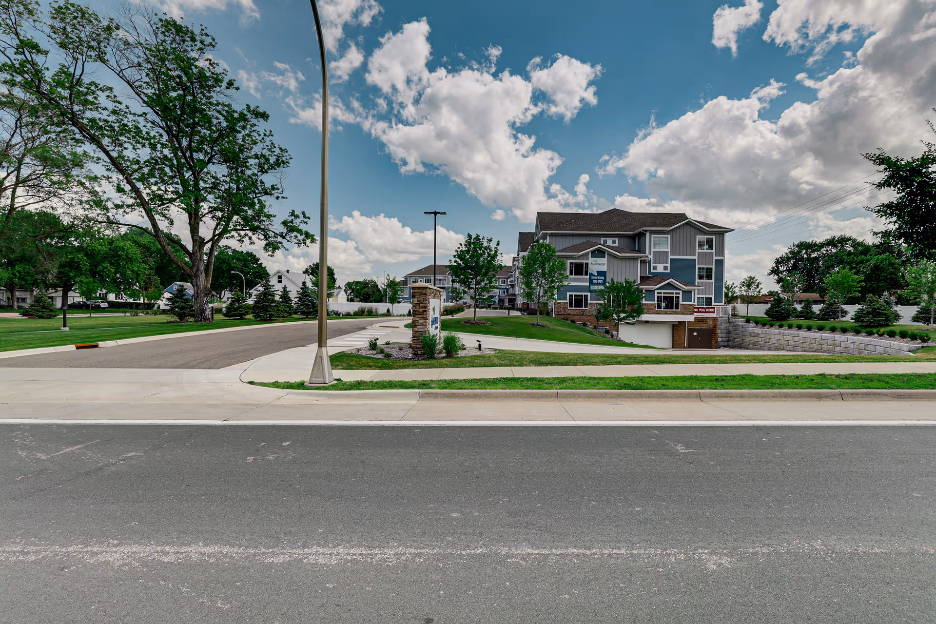 Exterior view of a senior living facility named Havenwood of Richfield, showing a multi-story building with gray siding and white trim, surrounded by green lawns, trees, and a clear blue sky with clouds.