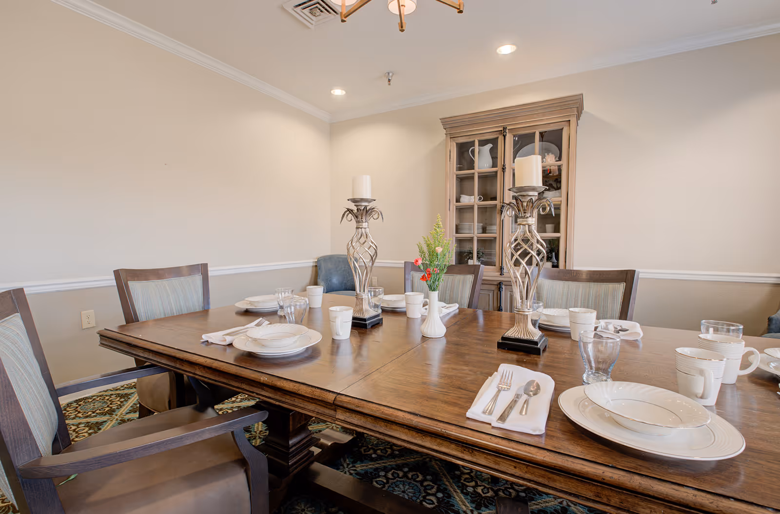 A formal dining room with a wooden table set for a meal, chairs, decorative candlesticks, and a china cabinet against a beige wall.