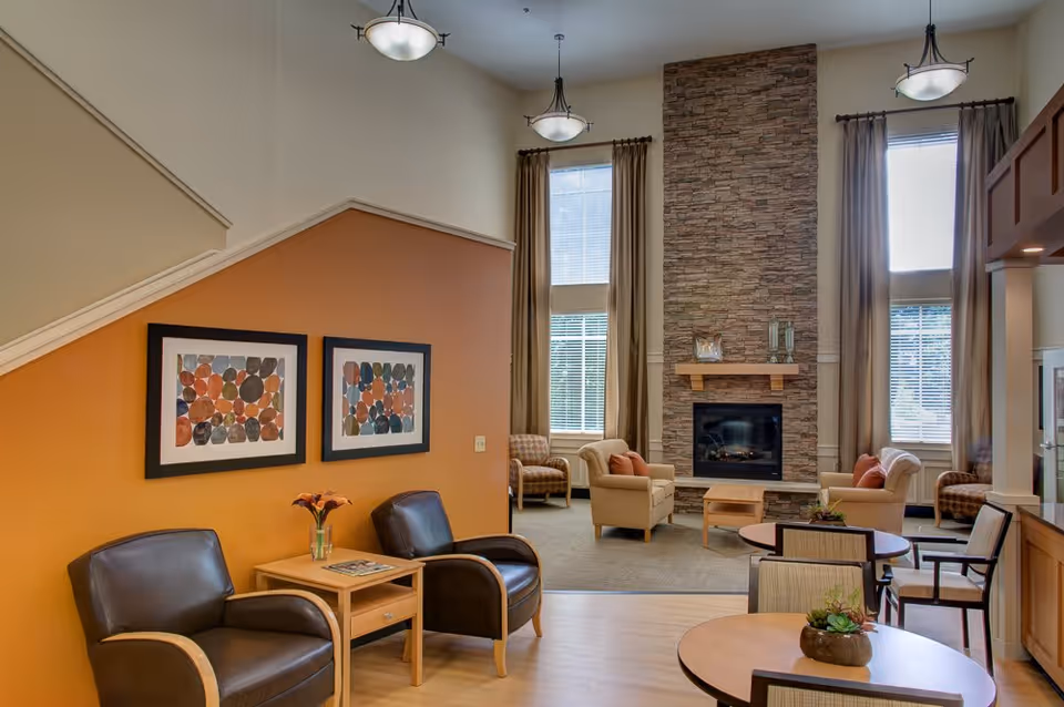 A spacious and well-lit living room area with high ceilings, featuring a stone fireplace flanked by tall windows with beige curtains. The room includes comfortable armchairs with orange cushions, a small wooden coffee table, round tables with chairs, and two leather chairs with a wooden side table against an orange accent wall adorned with two framed abstract artworks.