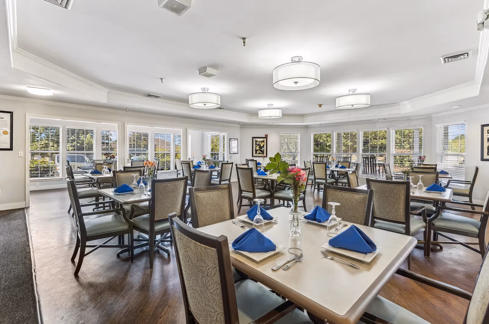 A bright and spacious dining room with multiple tables set for a meal. Each table has blue folded napkins, glassware, silverware, and small flower vases. Large windows with white blinds allow natural light to fill the room, and modern ceiling lights hang from the white ceiling. The room has wooden floors and comfortable chairs with patterned upholstery.