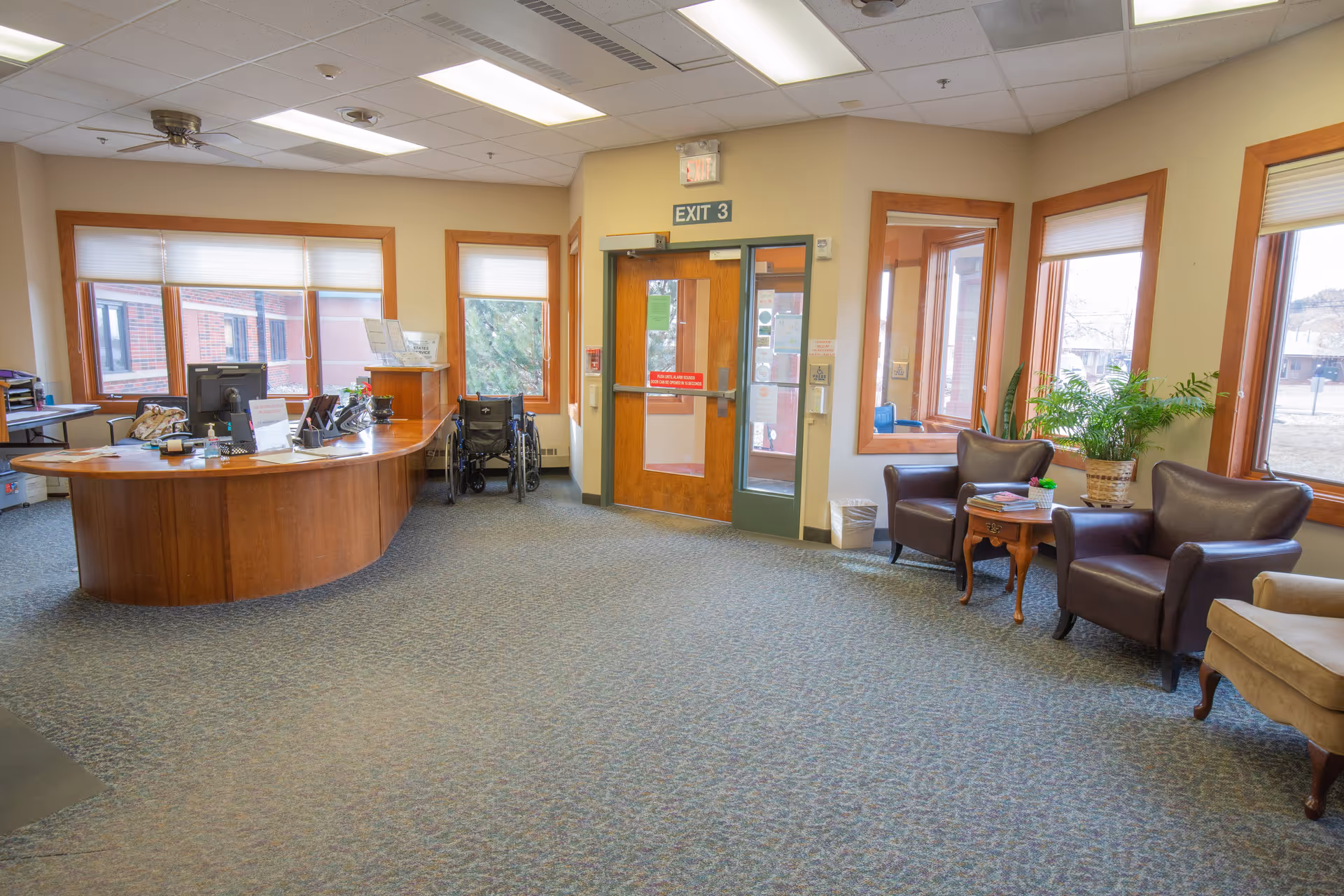 Reception area of Spearfish Canyon Healthcare with a curved wooden desk, computer, and office supplies. There is a wheelchair near the door labeled EXIT 3. The room has several large windows with blinds, two brown leather armchairs, a small wooden table with magazines, a potted plant, and carpeted flooring.