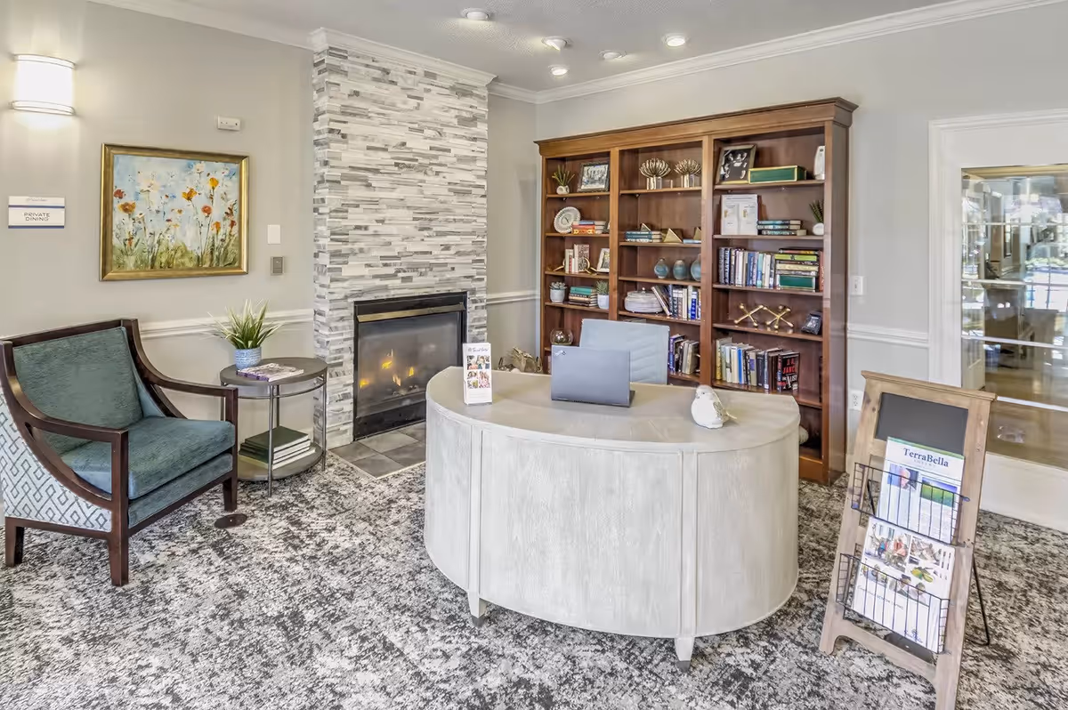 A cozy interior room with a modern stone fireplace, a wooden bookshelf filled with books and decorative items, a curved reception desk with a laptop and small decorative owl, a green upholstered armchair next to a small round table with a plant, and a wooden brochure stand displaying TerraBella brochures. The room has light-colored walls, a patterned carpet, and a painting of flowers on the wall.