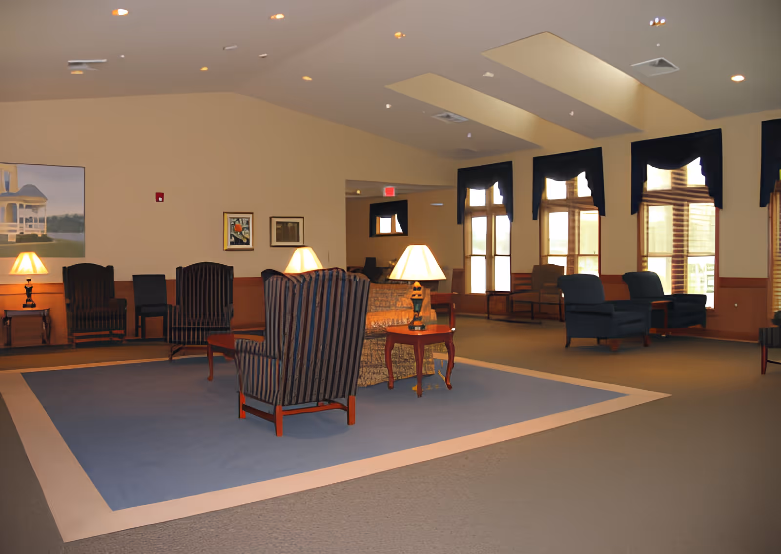A spacious common area in a senior living facility with multiple armchairs and sofas arranged around small wooden tables with lamps. The room has large windows with dark curtains, beige walls, and a high ceiling with recessed lighting. There are framed pictures on the walls and a blue area rug in the center.