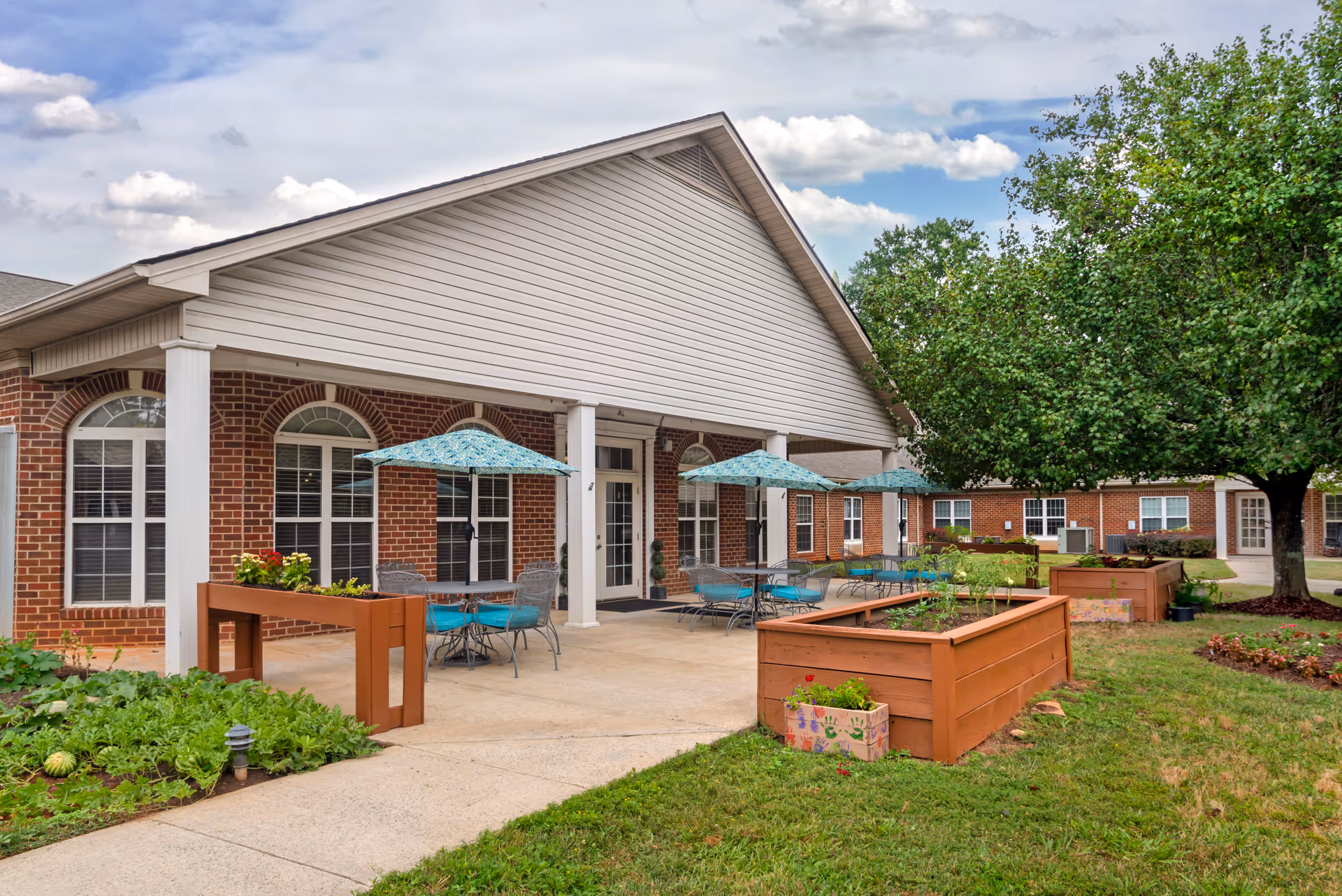 Brick senior living building with a covered patio, outdoor tables and umbrellas, and raised garden planters on a lawn.