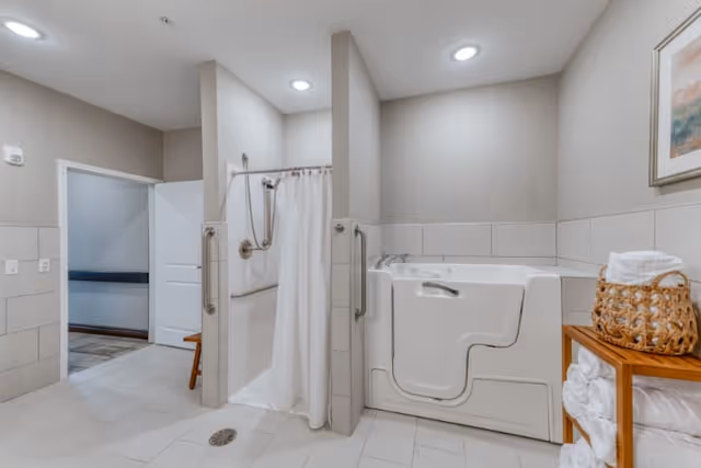 A senior-friendly bathroom featuring a walk-in shower with grab bars and a white curtain, alongside a walk-in bathtub with a door for easy access. The room has light-colored tiled floors and walls, recessed ceiling lights, and a wooden shelf holding neatly folded white towels and a basket. A framed artwork hangs on the wall above the shelf.
