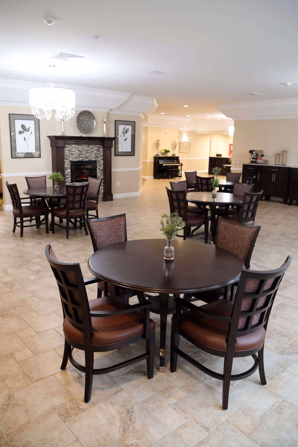 A spacious dining area with multiple round tables and chairs arranged neatly. Each table has a small vase with greenery. The room features a fireplace with decorative artwork above it, a chandelier, and a piano in the background. The floor is tiled and the walls are painted beige with white trim.