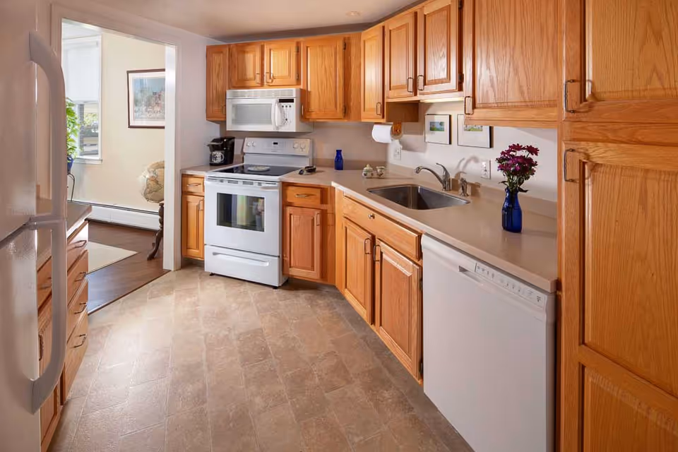 Bright kitchen with oak cabinets, white appliances, a sink and dishwasher, and a doorway to an adjacent living area.