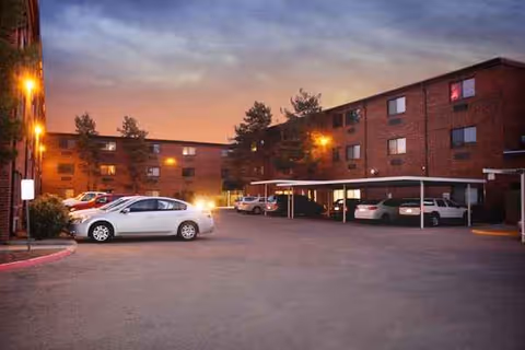 Brick senior living building with parked cars and a covered carport at dusk.