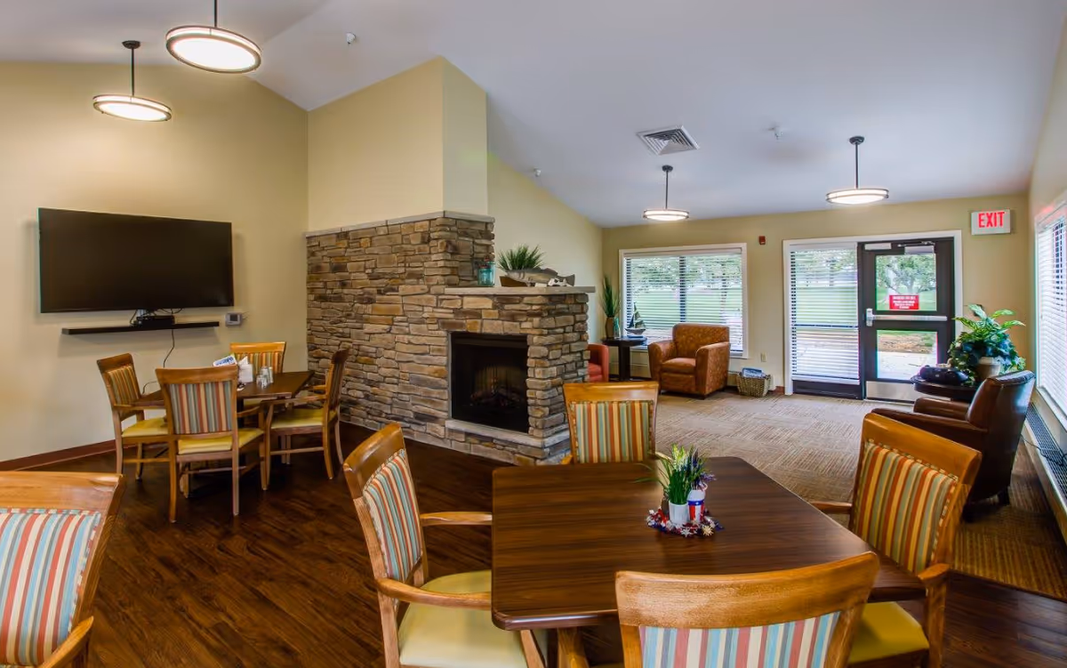 A senior living facility common area with wooden tables and striped cushioned chairs, a stone fireplace in the center, a wall-mounted flat-screen TV, and large windows letting in natural light. There are also comfortable armchairs near the windows and an exit door visible in the background.