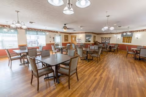 A spacious dining room with multiple square tables and cushioned chairs arranged neatly on a wooden floor. The room is well-lit with natural light coming through large windows covered with sheer curtains. Ceiling fans and hanging light fixtures provide additional lighting. The walls are decorated with framed pictures and a bulletin board.