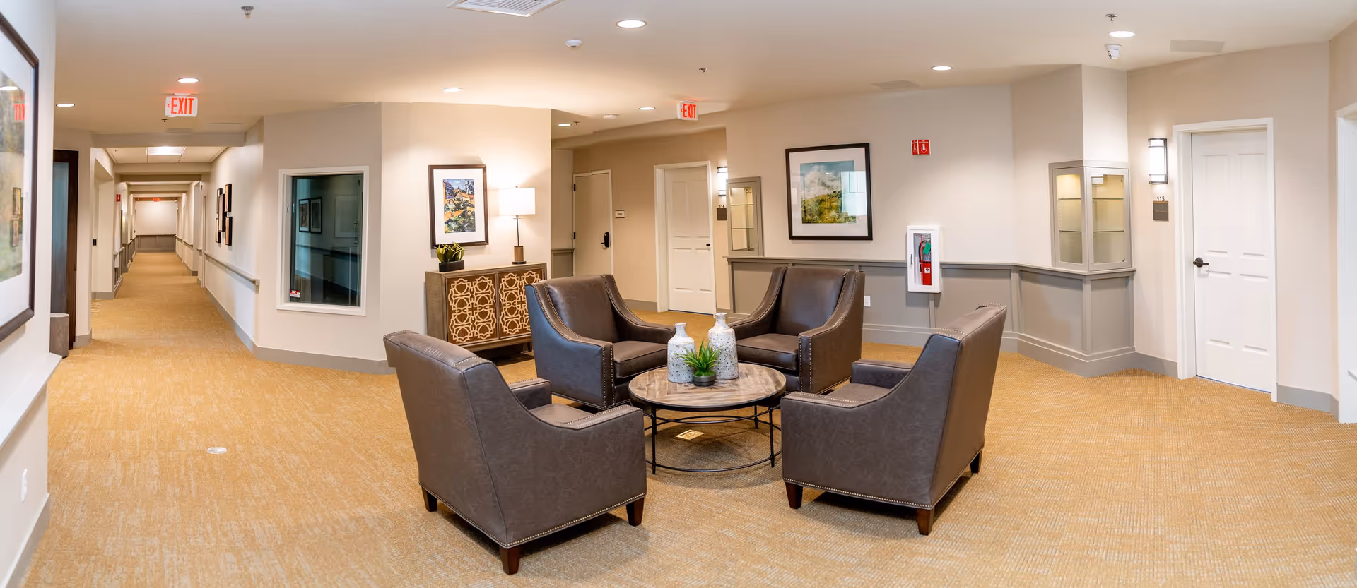A well-lit hallway in a senior living facility with beige walls and carpet. There is a seating area with four brown leather armchairs arranged around a round wooden coffee table with decorative vases and a small plant. The hallway has framed artwork on the walls, exit signs on the ceiling, and several doors leading to rooms. A fire extinguisher is mounted on the wall near one of the doors.