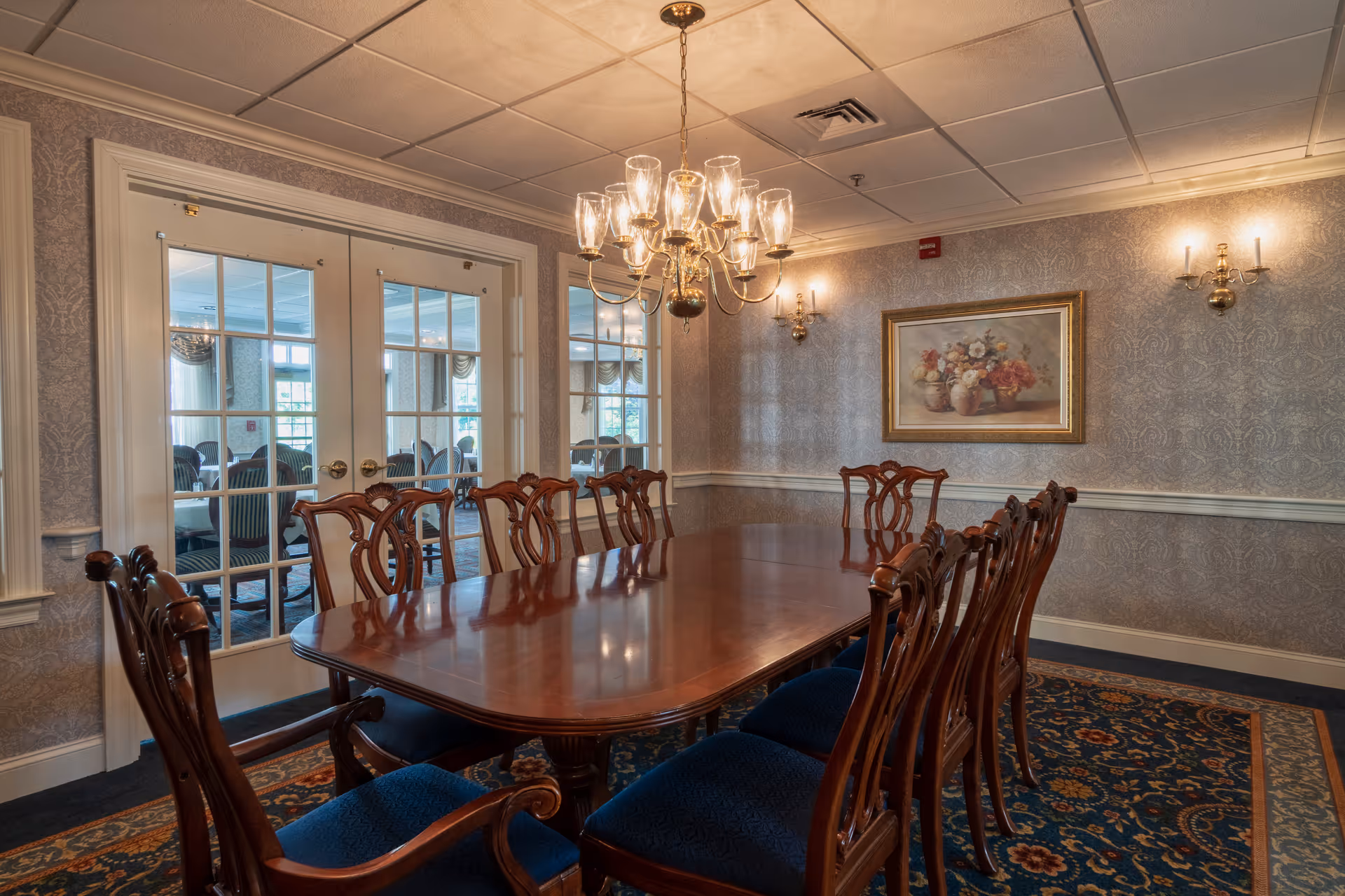 A formal dining room with a long polished wooden table surrounded by ten wooden chairs with blue upholstered seats. The room features patterned wallpaper, a decorative carpet, a chandelier with multiple glass shades hanging above the table, and wall sconces. French doors with glass panes lead to another room with additional seating.
