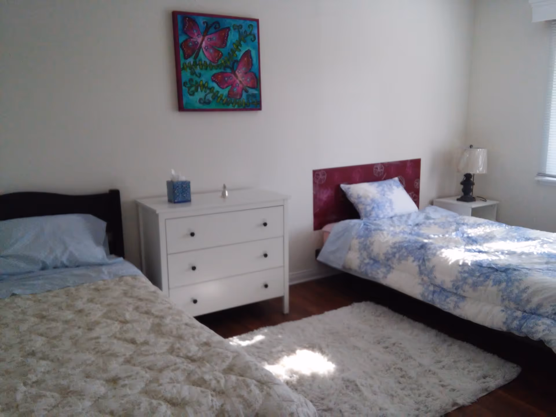 A bedroom with two single beds, one with a beige patterned blanket and the other with a blue and white floral blanket. Between the beds is a white three-drawer dresser with a tissue box and a small object on top. A colorful painting of two pink butterflies on a blue and green background hangs on the wall above the dresser. There is a white rug on the wooden floor and a window with blinds on the right side letting in natural light.
