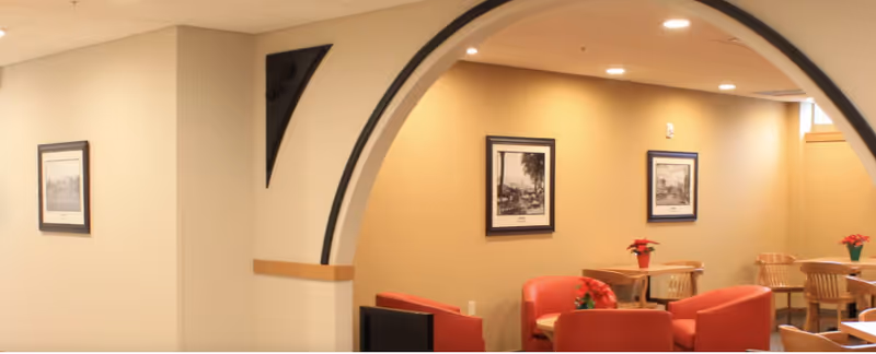 Interior common lounge/dining area with an arched opening, red armchairs, wooden tables, and framed pictures on warm-toned walls.
