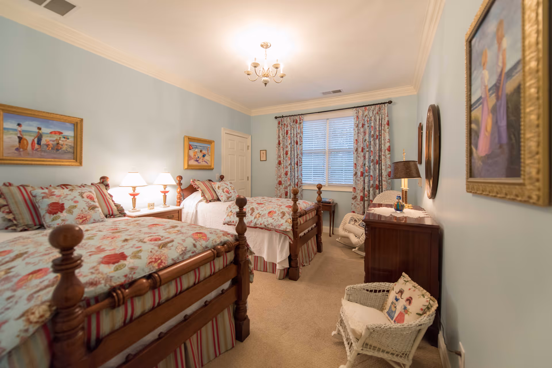 A cozy bedroom with two wooden twin beds featuring floral and striped bedding. The room has light blue walls adorned with framed paintings, a window with floral curtains, a wooden dresser with a lamp, and a white wicker chair with a decorative pillow. The room is softly lit by a ceiling chandelier and bedside lamps.