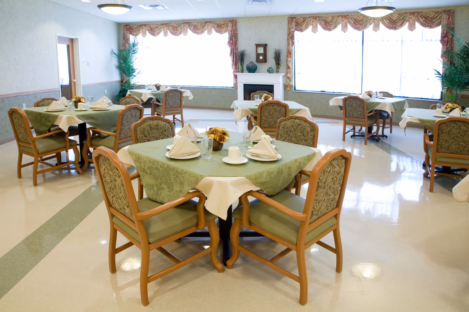 Dining room with several round tables covered in green tablecloths and chairs, each set with napkins and tableware.