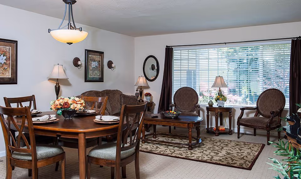 A cozy living and dining area with a wooden dining table set for four, a floral centerpiece, two upholstered armchairs, a coffee table, a sofa, table lamps, framed floral artwork on the walls, and a large window with blinds letting in natural light.