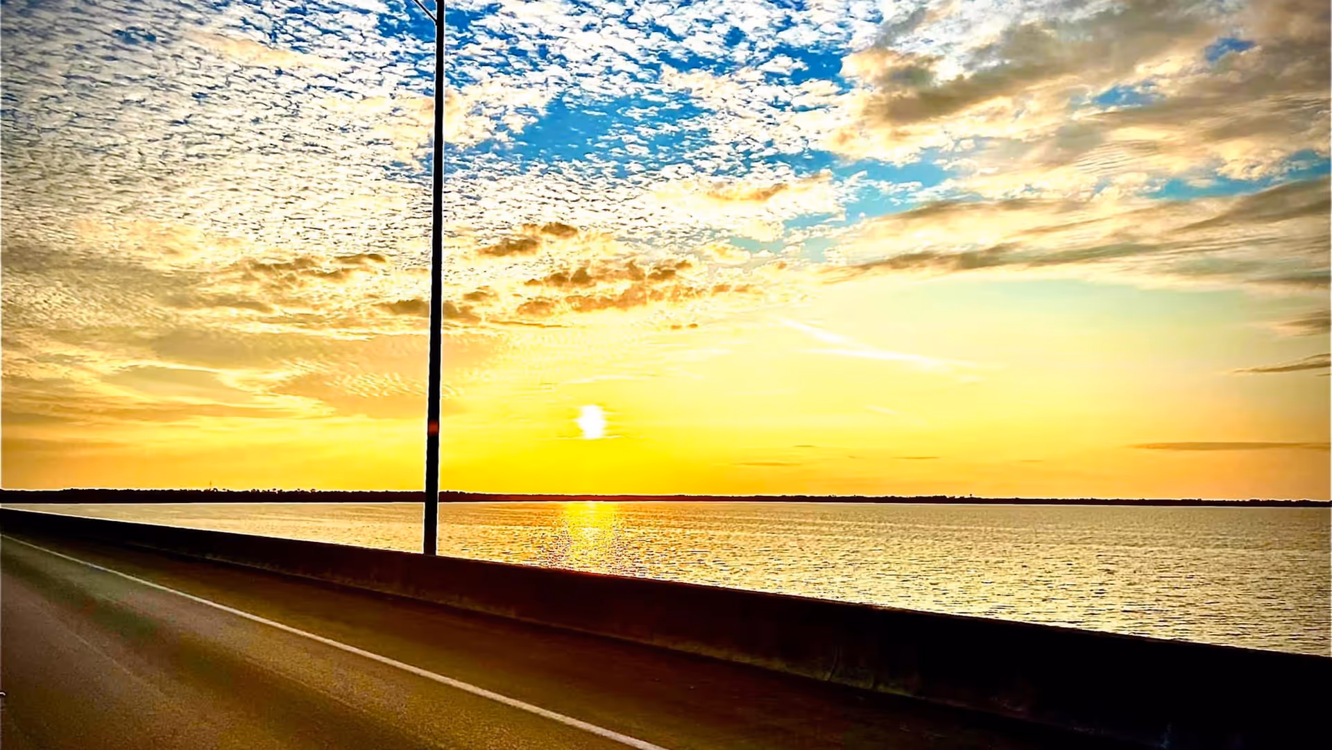 A scenic view of a sunset over a large body of water, seen from a road with a concrete barrier and a streetlight pole. The sky is partly cloudy with vibrant yellow and blue hues.