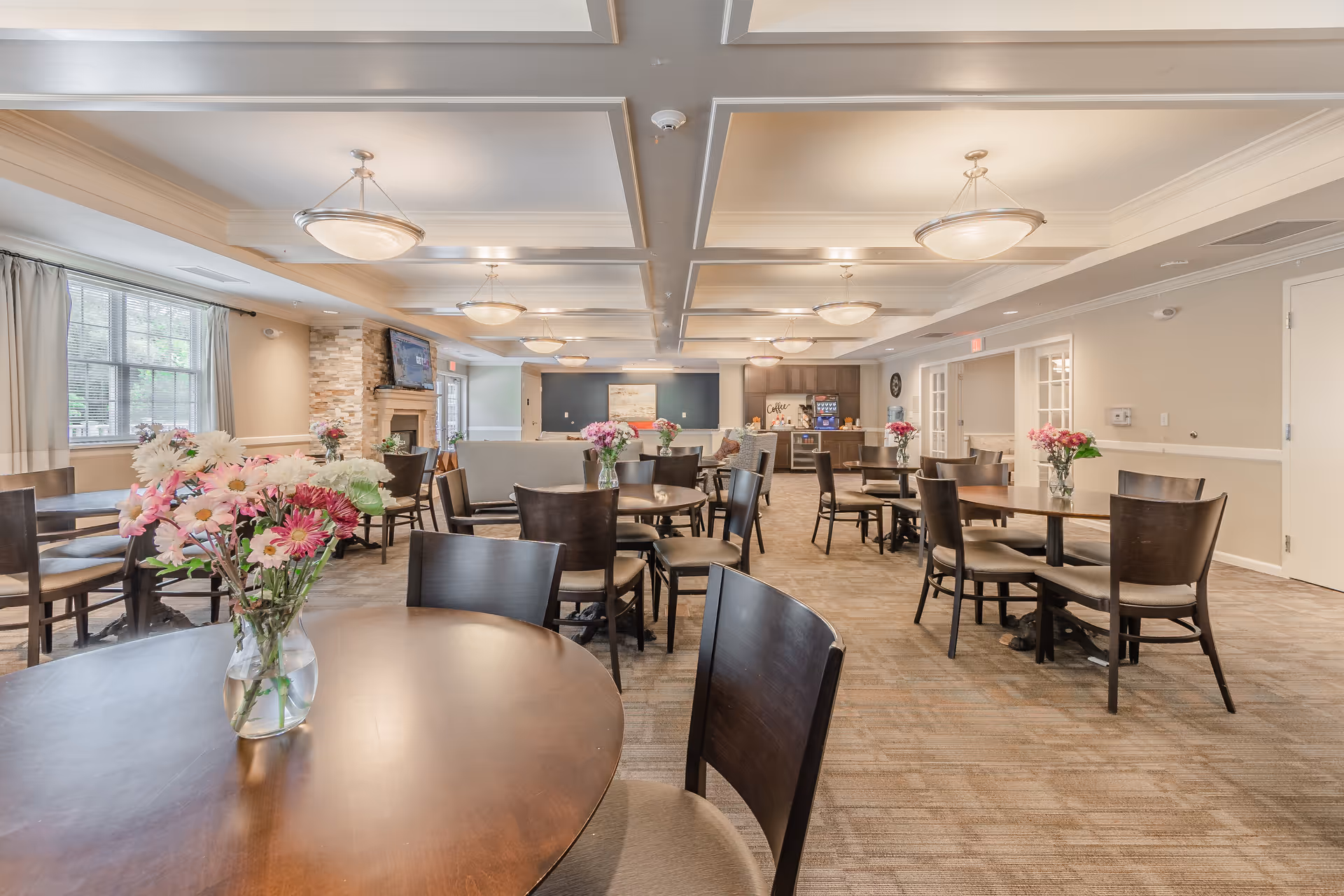 A spacious dining room with multiple round wooden tables and chairs, each table adorned with a vase of colorful flowers. The room features a coffered ceiling with hanging light fixtures, large windows with curtains, a stone fireplace with a mounted TV above it, and a coffee station in the background.