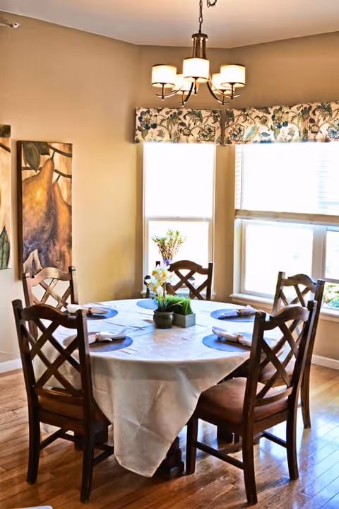 Bright dining room with a round table covered by a white tablecloth, six wooden chairs with place settings and a floral centerpiece beneath a chandelier by sunlit windows.