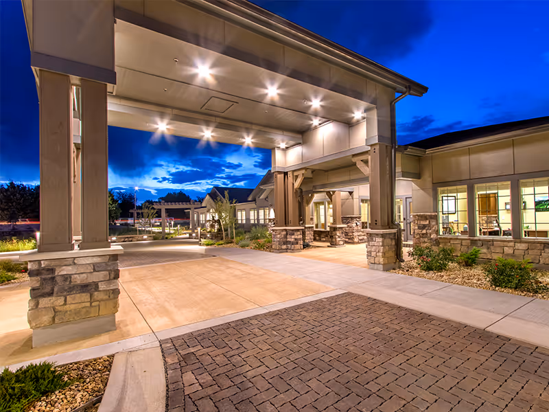 Entrance area of a senior living facility at dusk with a covered drop-off zone supported by stone and concrete pillars, illuminated by ceiling lights. The building has large windows and landscaped surroundings with shrubs and trees.