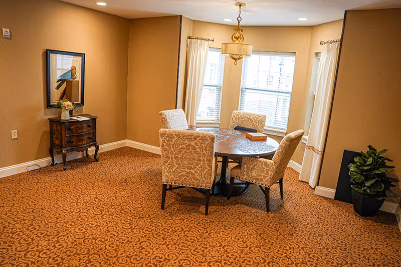 A cozy dining area with a round wooden table surrounded by four upholstered chairs with a floral pattern. The room has beige walls and carpet with a decorative pattern. There is a small wooden side table with a vase of flowers and a framed picture of a bird on the wall. Three windows with white curtains allow natural light into the room, and a gold chandelier hangs above the table. A potted plant is placed in the corner.